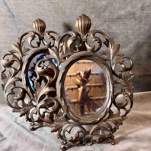 May include: Two antique brass oval picture frames with ornate, scrolling leaf and shell designs. One frame holds a small mirror, the other a blue and white ceramic piece. The frames stand on small feet.