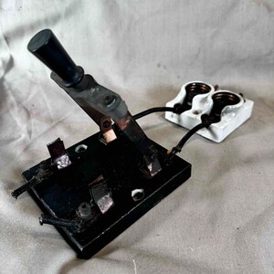 May include: A vintage black electrical switch with a lever handle and multiple copper contacts. The switch is connected to a white porcelain electrical outlet with two sockets.