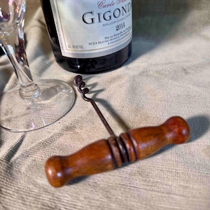 May include: A wooden-handled corkscrew with a metal spiral is positioned near a wine bottle and a wine glass. The bottle has a label that reads "GIGOND 2014". The corkscrew is made of wood and metal.