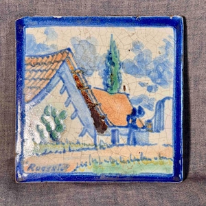 Puede incluir: Azulejo cerámico cuadrado con una escena de paisaje pintada a mano. La imagen presenta una casa con tejado naranja, un árbol alto verde y cielo azul. El azulejo está enmarcado con un borde azul oscuro y tiene la palabra "Rugezki" en la esquina inferior izquierda.