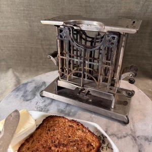 May include: An antique silver-toned toaster with intricate metalwork and wooden handles. A slice of toasted bread and a pat of butter sit on a small plate with a butter knife. The toaster is on a marble surface.