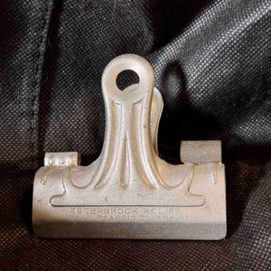 May include: A vintage silver-colored Esterbrook Relief Ball Bearing Clip No. 4. The clip has a rounded top with a hole and a rectangular base. The text "ESTERBROOK RELIEF BALL BEARING CLIP NO. 4" is visible.