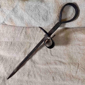 May include: An antique, dark metal candle holder. It features a long, pointed shaft, a circular handle, and a ring to catch wax. The candle holder is set against a neutral-toned, textured background.