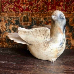 Puede incluir: Escultura de paloma de madera blanquecina desgastada, con las alas extendidas. El ave presenta un diseño detallado de plumas y un acabado ligeramente envejecido, lo que sugiere un estilo antiguo. La escultura se encuentra sobre un fondo estampado.
