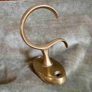 May include: A brass wall hook with a curved design. The hook is mounted on a round base with two screw holes.