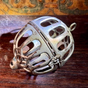 May include: An antique silver-colored metal cage-like object with an oval shape and openwork design. The object has a small loop at the top and a decorative clasp. The metal shows signs of age and wear, with a slightly tarnished appearance.