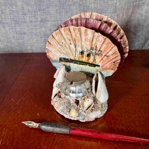 May include: A vintage seashell-themed inkwell with a ship painting, a clear glass ink pot, and a red-handled pen. The inkwell is decorated with various seashells and sand, creating a nautical aesthetic. The pen is positioned near the inkwell.