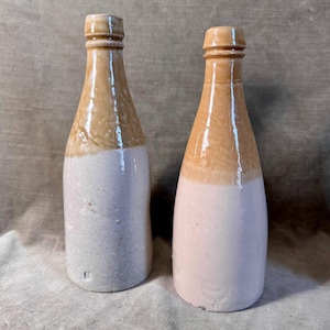 May include: Two antique stoneware bottles. The bottles have a two-tone design, with a light tan upper section and a white lower section. The bottles have a narrow neck and a rounded body. The bottles are likely vintage and suitable for display.