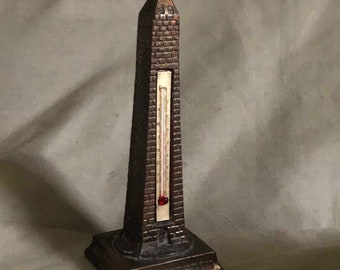 Könnte beinhalten: Ein vintage Thermometer aus Metall in Form des Washington Monuments. Das Thermometer hat eine rote Flüssigkeit im Inneren und ist mit Fahrenheit-Graden gekennzeichnet. Die Basis des Thermometers ist mit dem Wort "WASHINGTON" beschriftet.