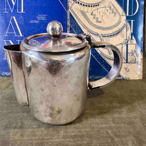 Könnte beinhalten: Eine silberne Teekanne mit einem Scharnierdeckel und einem gebogenen Griff. Die Teekanne steht auf einer grünen Oberfläche vor einem blau-weißen Hintergrund.