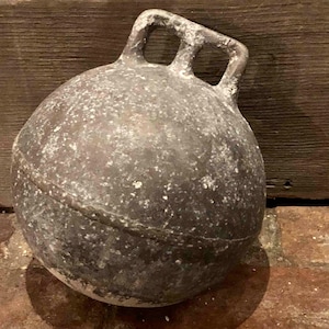 Puede incluir: Una boya de cerámica gris desgastada con dos bucles en la parte superior. La boya es redonda y tiene una textura rugosa.