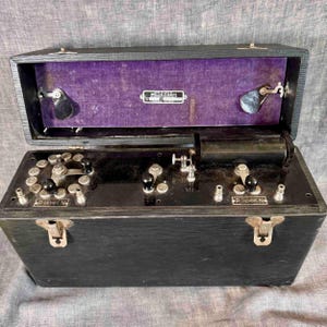 Puede incluir: Un antiguo dispositivo eléctrico negro McIntosh con tapa abisagrada, con varios diales, perillas y terminales. El interior está forrado con tela morada. El dispositivo tiene forma rectangular y cierres metálicos.
