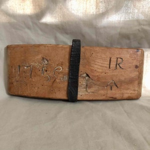 Op de afbeelding: Een houten bord met het getal 1756 erin gegraveerd, samen met de letters IR en een pijl die omhoog wijst. Het bord is bevestigd aan een zwarte leren riem.