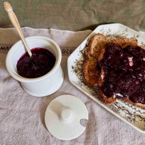 May include: A white ceramic jam pot with a silver spoon inside, filled with dark red jam. Beside it, a plate of toast covered in jam. A white lid with a handle sits nearby. The scene is set on a patterned cloth.