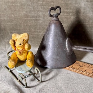 May include: A vintage scene featuring a small, yellow teddy bear seated in a miniature blue and green wheeled chair. Beside it is a large, cone-shaped metal object with a heart-shaped handle. A wooden ruler is also present.