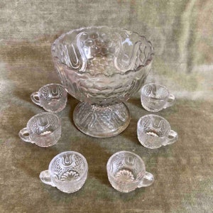 Puede incluir: Un juego de ponchera de cristal vintage con seis tazas. La ponchera y las tazas son de cristal transparente con un diseño texturizado. La ponchera tiene una base de pedestal.