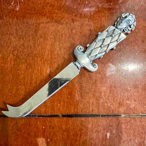 May include: A silver cheese knife with a decorative handle featuring a face and a diamond pattern. The blade has a serrated edge and a curved tip. The knife is designed for cutting cheese.