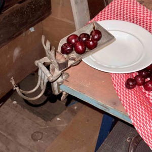 以下が含まれることがあります： 新鮮で熟したさくらんぼをすくう、ヴィンテージのさくらんぼの種取り器。白いお皿と赤と白のチェック柄の布が近くにあります。種取り器は木製の表面に取り付けられています。