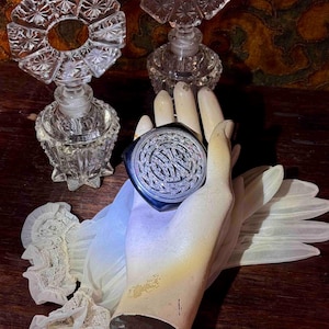 May include: A vintage compact mirror with a silver-tone filigree design, held by a mannequin hand, alongside two antique glass perfume bottles. The compact has a dark blue border. The background is a dark wood surface.