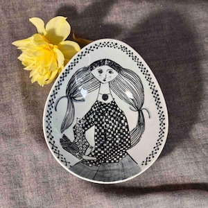 Puede incluir: Plato ovalado de cerámica con una ilustración en blanco y negro de una niña sosteniendo un pájaro. La niña tiene el pelo largo y lleva un vestido a cuadros. El plato tiene un borde decorativo y está junto a un narciso amarillo.
