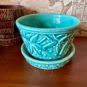 Könnte beinhalten: Ein türkisfarbener Keramik-Blumentopf mit einem Blattdesign und einer passenden Untertasse. Der Topf hat eine strukturierte Oberfläche und einen leicht ausgestellten Rand.