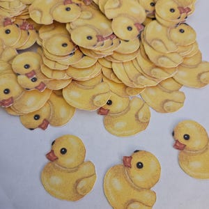 May include: A collection of yellow duck-shaped paper cutouts with orange beaks and black eyes. The cutouts are scattered, with several in focus, showcasing the detailed illustration of the ducks. The overall theme is a craft or decorative item.