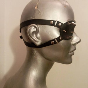 May include: Black leather eye mask with silver metal studs and straps. The mask features a central eye covering and adjustable straps. The studs are evenly spaced across the mask's surface.