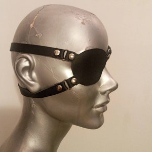 Könnte beinhalten: Schwarze Augenmaske aus Leder mit verstellbaren Riemen und silberfarbenen Metallbeschlägen. Die Maske bedeckt ein Auge und wird mit einem Riemen um den Kopf befestigt. Die Augenmaske wird auf einem Schaufensterkopf gezeigt.