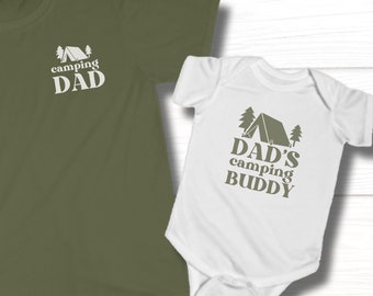 May include: A green t-shirt with the text "camping DAD" and a camping tent graphic. A white onesie with the text "DAD'S camping BUDDY" and a camping tent graphic.
