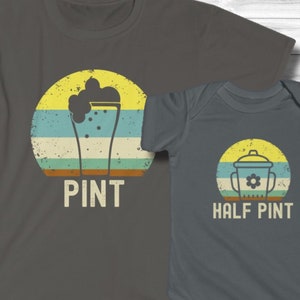 May include: Two gray t-shirts with matching designs. One says "PINT" with a beer glass graphic inside a retro sunset. The other is a baby onesie that says "HALF PINT" with a similar graphic of a baby bottle.
