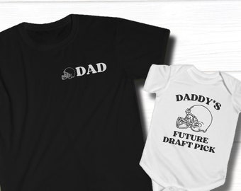 Puede incluir: Una camiseta negra con la palabra "DAD" y un gráfico de casco de fútbol. Un body blanco con el texto "DADDY'S FUTURE DRAFT PICK" y un gráfico de casco de fútbol.
