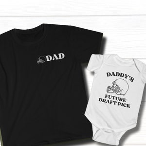 Puede incluir: Una camiseta negra con la palabra "DAD" y un gráfico de casco de fútbol. Un body blanco con el texto "DADDY'S FUTURE DRAFT PICK" y un gráfico de casco de fútbol.