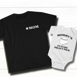 Puede incluir: Una camiseta negra con una pelota de béisbol y la palabra "MOM" impresa en ella. Un mameluco blanco con una pelota de béisbol y el texto "Mommy's Future Draft Pick" impreso en él.