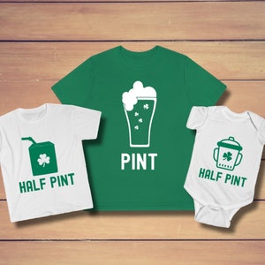 Può includere: Tre magliette bianche con disegni di trifoglio verde. La maglietta più grande dice "PINT" e ha un'immagine di un bicchiere di birra. La maglietta media dice "HALF PINT" e ha un'immagine di una scatola di succo. La maglietta più piccola dice "HALF PINT" e ha un'immagine di un biberon.