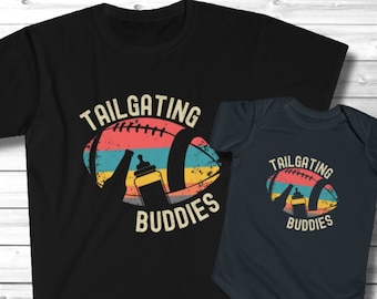 Puede incluir: Una camiseta negra y un body de bebé negro con un gráfico de fútbol de estilo retro y el texto "Tailgating Buddies". El gráfico de fútbol presenta un biberón dentro del balón de fútbol.