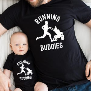 Könnte beinhalten: Schwarzes T-Shirt und Baby-Body-Set. Beide Artikel zeigen den Text "RUNNING BUDDIES" und eine weiße Grafik einer Person, die mit einem Kinderwagen läuft. Das Baby trägt den Body, und der Erwachsene trägt das T-Shirt.