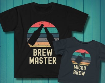 Könnte beinhalten: Zwei schwarze T-Shirts mit einem Retro-Sonnenuntergangsdesign. Das größere Shirt trägt den Schriftzug "Brew Master" und zeigt zwei anstoßende Bierflaschen. Das kleinere Shirt trägt den Schriftzug "Micro Brew" und zeigt eine Babyflasche.