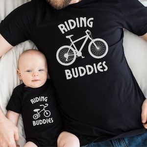 Peut inclure: Un t-shirt noir avec le texte "Riding Buddies" et un graphique de vélo. Un bébé porte un body noir avec le texte "Riding Buddies" et un graphique de tricycle.