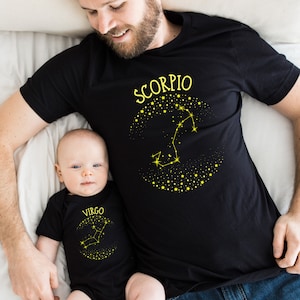 May include: A black t-shirt with the word "Scorpio" and a constellation design in yellow. A baby is wearing a black onesie with the word "Virgo" and a constellation design in yellow.
