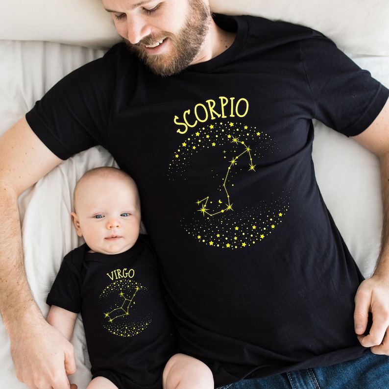May include: A black t-shirt with the word "Scorpio" and a constellation design in yellow. A baby is wearing a black onesie with the word "Virgo" and a constellation design in yellow.
