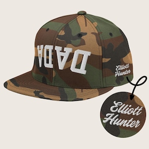 May include: A camouflage-patterned baseball cap with a white embroidered "DADA" on the front and "Elliott Hunter" embroidered on the side.