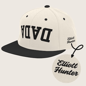 May include: A white and black baseball cap with the word "DADA" embroidered in black on the front. The cap has a black brim and a small embroidered patch on the side that says "Elliott Hunter".