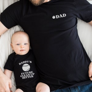 Puede incluir: Un padre y un hijo vistiendo camisetas negras a juego con texto de temática de béisbol. La camiseta del padre dice "DAD" con un gráfico de béisbol. La camiseta del hijo dice "Daddy's Future Clean Up Hitter" con un gráfico de béisbol.