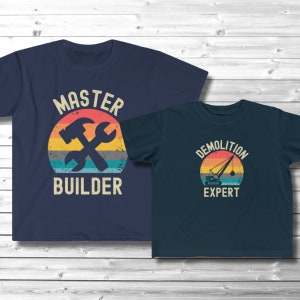 Op de afbeelding: Twee marineblauwe T-shirts met een grafisch ontwerp in vintage stijl. Het grotere shirt zegt "Master Builder" met een hamer en een moersleutel in een cirkel. Het kleinere shirt zegt "Demolition Expert" met een kraan in een cirkel.