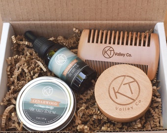 May include: A beard care kit in a cardboard box containing a 1 oz bottle of Cedarwood beard oil, a 2 oz tin of Cedarwood beard balm, and a wooden comb. Each item features the KT Valley Co. logo, with the balm and oil labels clearly visible.