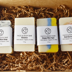 May include: Four bars of soap in a brown box with shredded paper. The soap bars are white with different coloured swirls and have labels that say "Face & Body Bar" with different scents: "Natural Man Soap", "Balance", "Happy Men Type", and "Oak Barrel Cider".