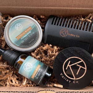Könnte beinhalten: Eine Geschenkbox gefüllt mit Bartpflegeprodukten, darunter ein Sandholz-Bartbalsam, Bartöl und ein Holzkamm. Die Box ist mit braunen Papierschnitzeln ausgekleidet. Die Produkte sind mit dem Text "Sandalwood Premium Quality Beard Balm" und "Sandalwood Premium Quality Beard Oil" gekennzeichnet. Der Kamm ist mit dem Text "K Valley Co." beschriftet.