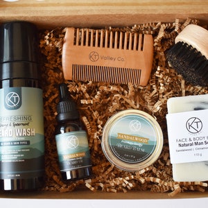 May include: A gift box set for beard care, containing a beard wash, beard oil, beard balm, a wooden comb, a wooden brush, and a bar of soap. The products are all in a brown cardboard box with shredded paper.