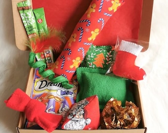 May include: A cardboard box filled with Christmas-themed cat toys. The toys include a red and white catnip toy shaped like a stocking, a green and white catnip toy shaped like a Christmas tree, a red and white catnip toy shaped like a gingerbread man, a green catnip toy shaped like a pillow, a red and white catnip toy shaped like a Santa Claus, and a gold foil catnip toy. The box also includes a package of Dreamies cat treats.