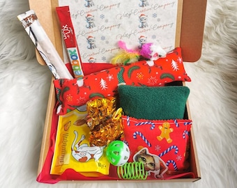 May include: A festive cat gift box filled with toys and treats. The box contains red and green patterned cat toys, a green plush toy, a small ball, and a spring toy. The box is lined with paper featuring a repeating pattern of cats wearing Santa hats.